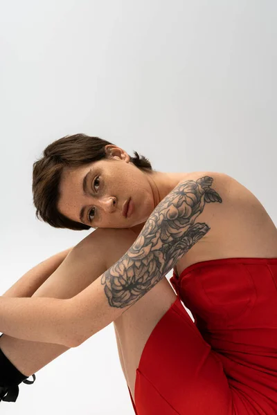 Мечтательная татуированная женщина в красном корсетном платье сидит и смотрит в камеру на сером фоне — стоковое фото