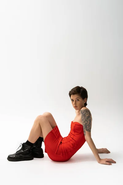 Молодая татуированная женщина в черных грубых сапогах и красном корсете платье сидит и смотрит на камеру на сером фоне — стоковое фото
