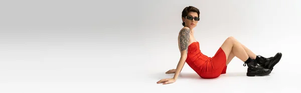Full length of slim tattooed woman in black boots and red corset dress sitting on grey background, banner — Stock Photo