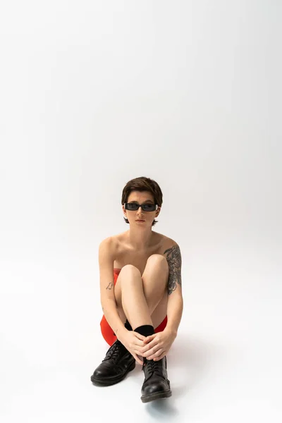 Full length of young tattooed woman in black rough boots and trendy sunglasses sitting on grey background — Stock Photo