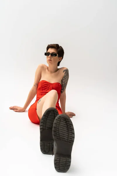 Повна довжина модної жінки в червоному корсетному платті і сонцезахисних окулярах, сидячи і дивлячись на сірий фон — стокове фото