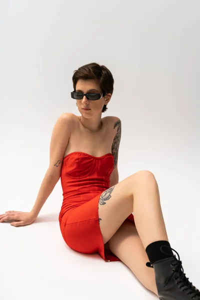 Brunette tattooed woman in sunglasses and red corset dress sitting on grey background — Stock Photo