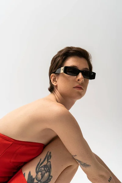 Mujer tatuada en gafas de sol con estilo y vestido rojo sin tirantes mirando a la cámara aislada en gris - foto de stock