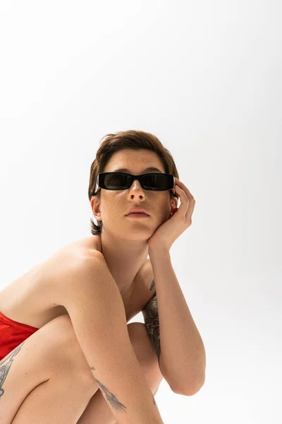 Молодая татуированная женщина с короткими волосами брюнетки позирует в стильных солнцезащитных очках на сером фоне — стоковое фото