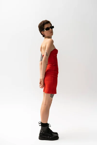 Piena lunghezza della donna sottile tatuato in occhiali da sole e rosso abito senza spalline con stivali neri su sfondo grigio — Foto stock
