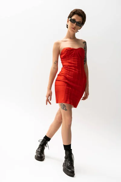 Повна довжина татуйованої жінки в червоній сукні та чорних шкіряних чоботях на сірому фоні — стокове фото