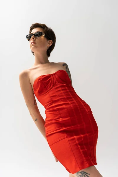 Visão de baixo ângulo da mulher esbelta em vestido sem alças vermelho e óculos de sol elegantes no fundo cinza — Fotografia de Stock