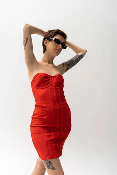 Low angle view of tattooed woman in sunglasses and red corset dress posing with hands behind head on grey — Stock Photo
