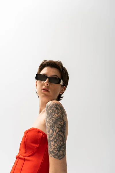Brunette tattooed woman in sunglasses and red corset dress looking at camera on grey background — Stock Photo