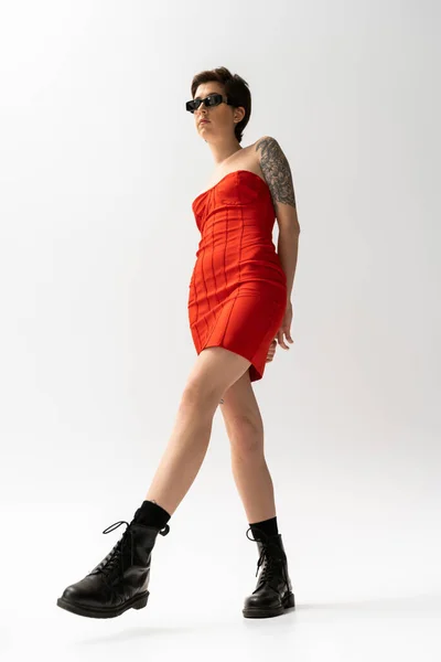 Volle Länge der schlanken tätowierten Frau posiert in rotem Korsettkleid und schwarzen Stiefeln auf grauem Hintergrund — Stockfoto