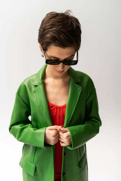 Jeune femme brune posant dans des lunettes de soleil à la mode et veste en cuir vert sur fond gris — Photo de stock