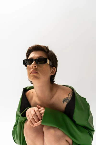 Татуированная женщина в зеленой кожаной куртке и модных солнцезащитных очках сидит на сером фоне — стоковое фото