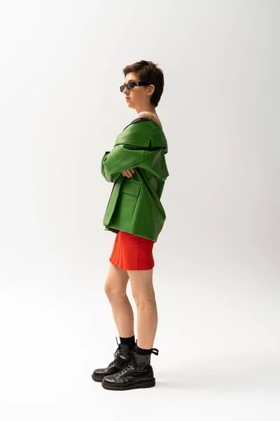 Longitud completa de la mujer con estilo en chaqueta de cuero verde y botas negras de pie con los brazos cruzados sobre fondo gris - foto de stock