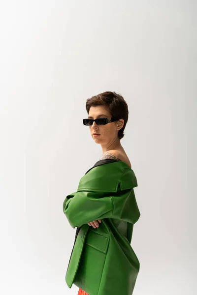 Brünett tätowierte Frau in grüner Lederjacke und stylischer Sonnenbrille blickt vereinzelt in die Kamera auf grau — Stockfoto