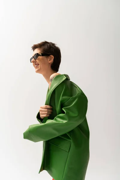 Side view of overjoyed brunette woman posing in trendy sunglasses and green leather jacket isolated on grey — Stock Photo