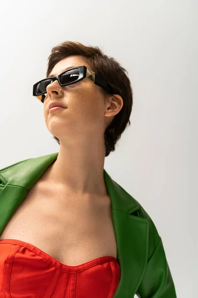 Low angle view of young brunette woman in sunglasses and green leather jacket looking away isolated on grey — Stock Photo