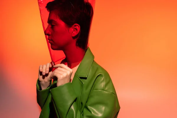 Seitenansicht der trendigen Frau in grüner Lederjacke und transparentem Kunststoff in Zylinderform auf orangefarbenem und pinkem Hintergrund — Stockfoto