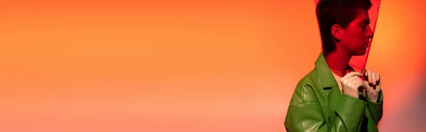 Vue latérale de la jeune femme brune en veste en cuir vert et plastique transparent sur la tête sur fond orange et rose, bannière — Photo de stock