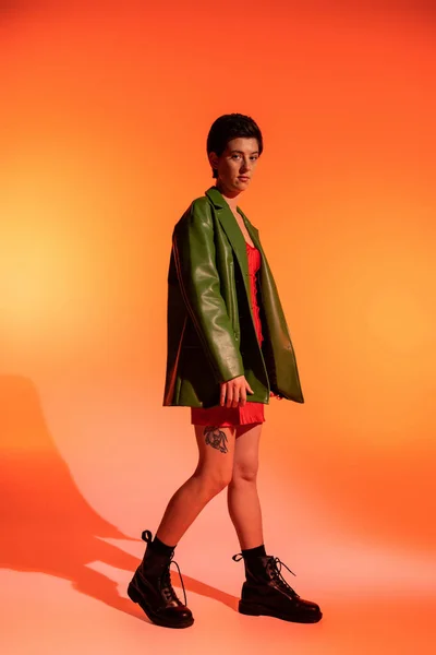 Full length of tattooed woman in green leather jacket and black boots looking at camera on orange background — Stock Photo