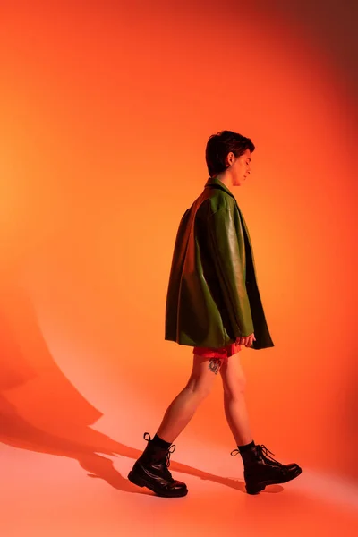 Vista lateral de mujer tatuada en chaqueta verde y botas negras caminando sobre fondo naranja - foto de stock