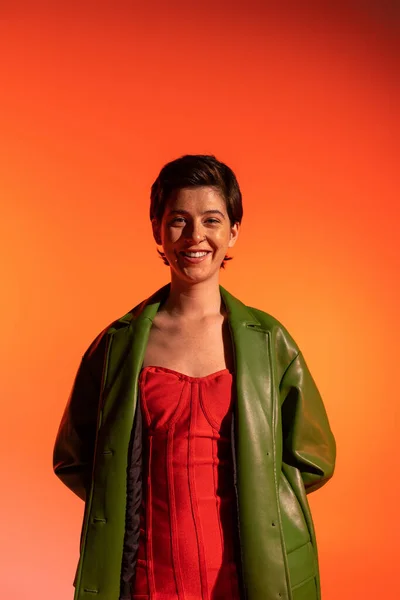 Donna bruna spensierata in giacca di pelle verde e abito rosso corsetto guardando la fotocamera su sfondo arancione — Foto stock