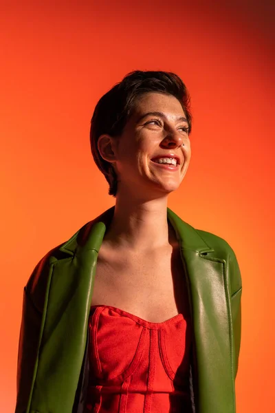 Fröhliche brünette Frau in rotem Korsettkleid und grüner Lederjacke lächelt und schaut auf orangefarbenem Hintergrund weg — Stockfoto
