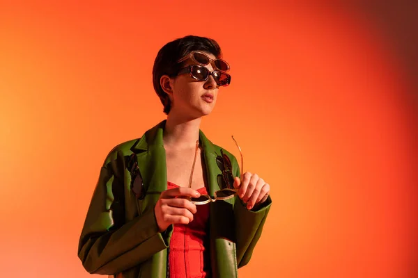 Mulher na moda em jaqueta de couro verde posando em vários óculos de sol e olhando para o fundo laranja — Fotografia de Stock