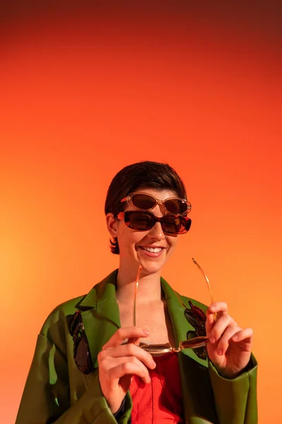 Alegre morena mujer con chaqueta de cuero verde y varias gafas de sol con estilo sobre fondo naranja - foto de stock
