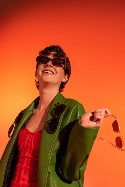 Збуджена і модна жінка в зеленій шкіряній куртці позує з різними сонцезахисними окулярами на помаранчевому фоні — стокове фото