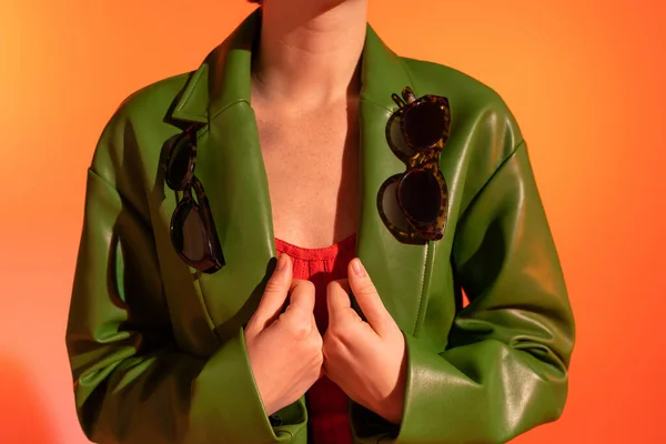 Visão parcial da mulher com óculos de sol elegantes na jaqueta de couro verde no fundo laranja — Fotografia de Stock