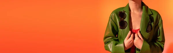 Vista ritagliata della donna in posa con occhiali da sole alla moda su giacca di pelle verde su sfondo arancione, banner — Foto stock