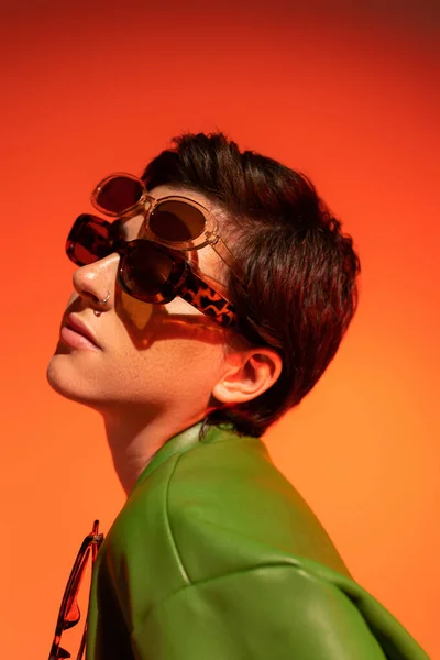 Portrait of young woman with short brunette hair wearing two trendy sunglasses on orange background — Stock Photo
