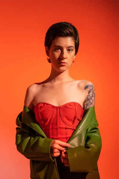 Joven mujer tatuada en vestido de corsé rojo y chaqueta de cuero verde mirando a la cámara sobre fondo naranja - foto de stock