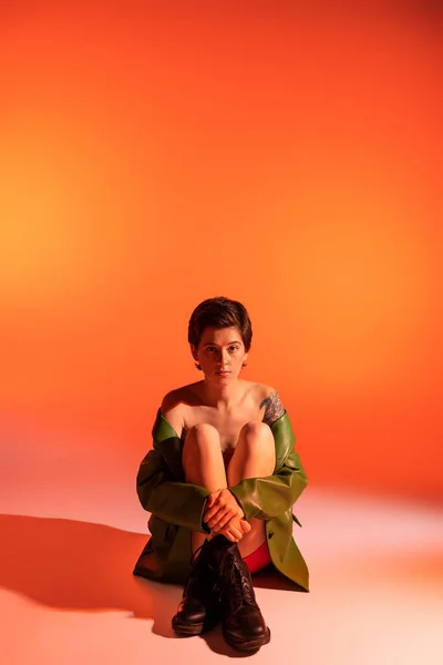 Pleine longueur de femme tatouée brune en bottes rugueuses noires et veste verte assise sur fond orange — Photo de stock