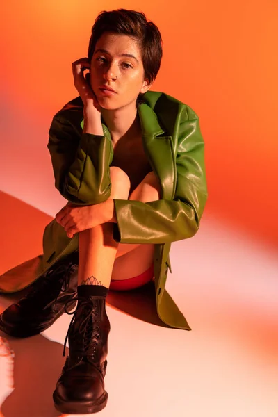 Повна довжина мрійливої жінки в зеленій шкіряній куртці і чорних чоботях, сидячи і дивлячись на камеру на помаранчевому фоні — стокове фото
