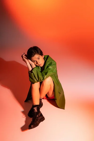 Модная и задумчивая женщина в черных кожаных сапогах и зеленой куртке сидит и смотрит в камеру на красочном фоне — стоковое фото