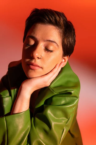 Porträt einer brünetten Frau in grüner Lederjacke, die mit geschlossenen Augen und händennahem Gesicht auf orangefarbenem und rosa Hintergrund posiert — Stockfoto