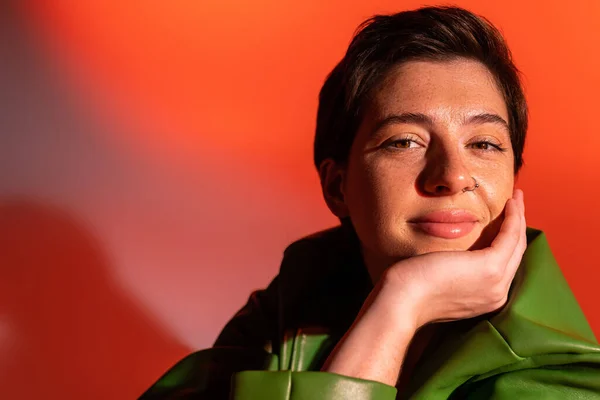 Portrait of cheerful young woman in green jacket holding hand near face and smiling at camera on colorful background — Stock Photo