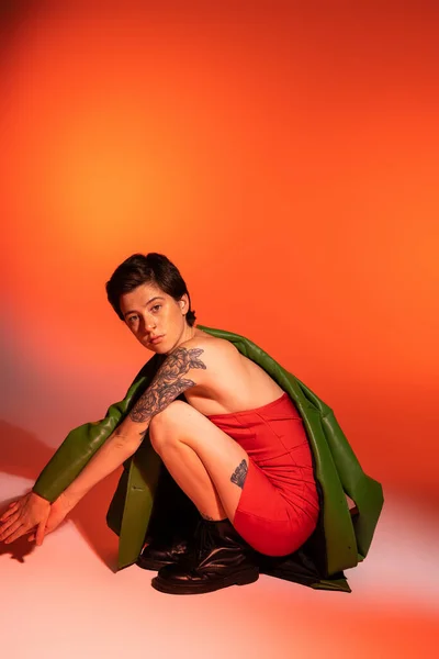 Comprimento total de mulher tatuada em vestido de espartilho vermelho e botas pretas sentadas em assombrações e olhando para a câmera em fundo laranja e rosa — Fotografia de Stock