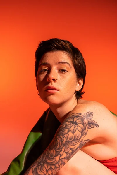 Портрет задумчивой женщины с татуировкой и короткими волосами брюнетки, смотрящей в камеру на оранжевом фоне — стоковое фото