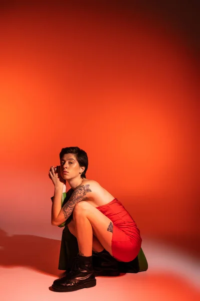 Trendy tattooed woman in red strapless dress and black leather boots posing on haunches on orange background — Stock Photo