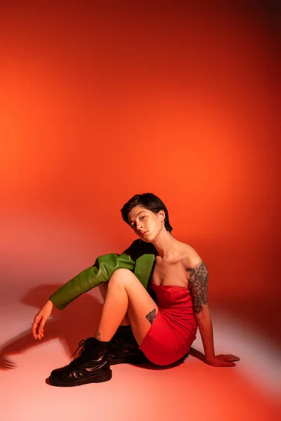 Trendige tätowierte Frau in rotem trägerlosem Kleid und schwarzen Stiefeln blickt in die Kamera, während sie auf orangefarbenem Hintergrund posiert — Stockfoto