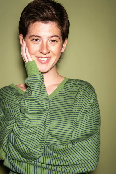 Ritratto di donna bruna gioiosa in pullover a righe che tocca il viso e guarda la fotocamera su sfondo verde — Foto stock