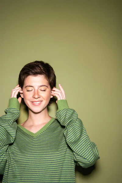Young and stylish woman with closed eyes fixing short brunette hair on green background — Stock Photo
