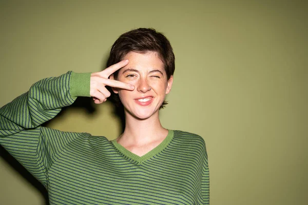 Cheerful woman in striped jumper showing victory sign near face and winking at camera on green background — Stock Photo