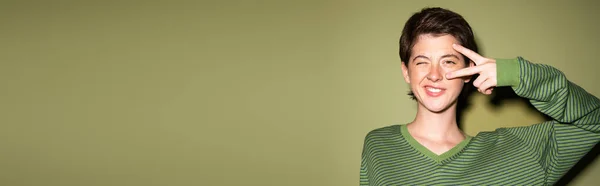 Donna bruna sorridente strizzando l'occhio alla macchina fotografica e mostrando il segno della pace vicino al viso su sfondo verde, banner — Foto stock