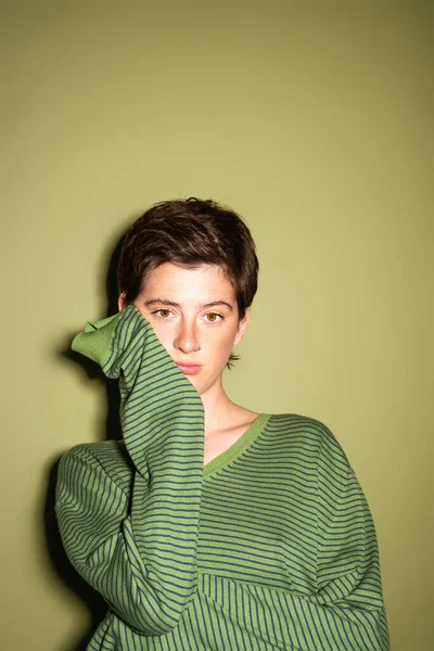 Задумчивая брюнетка в модном пуловере смотрит в камеру на зеленом фоне — стоковое фото