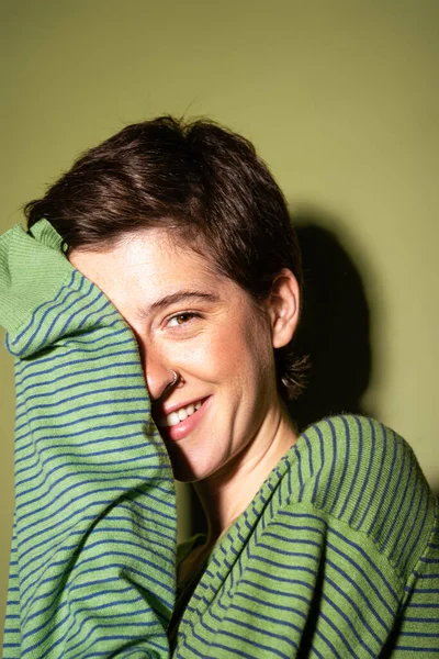 Happy brunette woman looking at camera and obscuring face with striped jumper on green background — Stock Photo