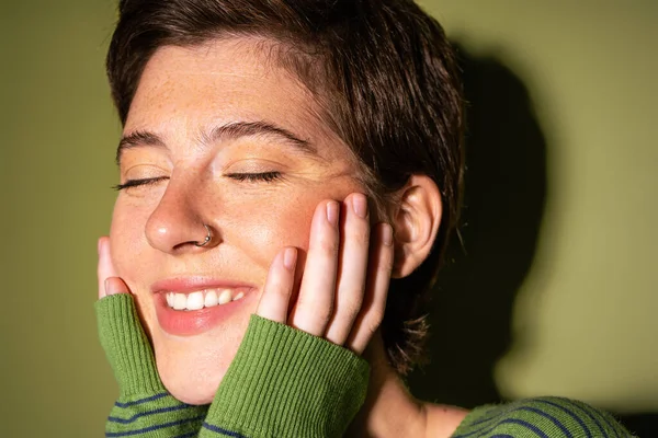Retrato de mulher radiante com sardas e nariz perfurante tocando o rosto e sorrindo com os olhos fechados no fundo verde — Fotografia de Stock