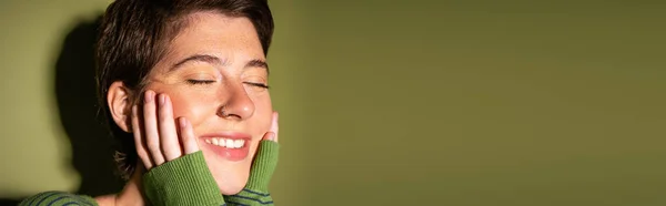 Retrato de mujer pecosa despreocupada sonriendo con los ojos cerrados y sosteniendo las manos cerca de la cara sobre fondo verde, bandera - foto de stock